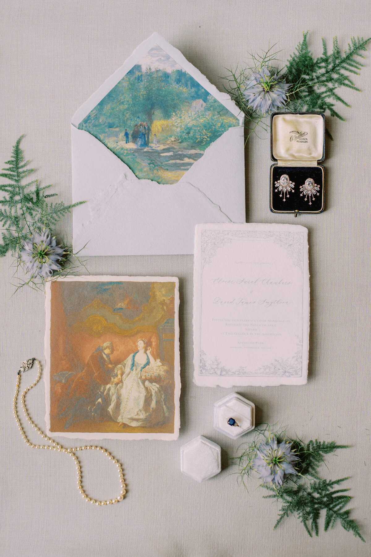 flatlay of a blue vintage wedding stationery set on natural paper displayed with a sapphire engagement ring and pearl necklace and blue flower heads