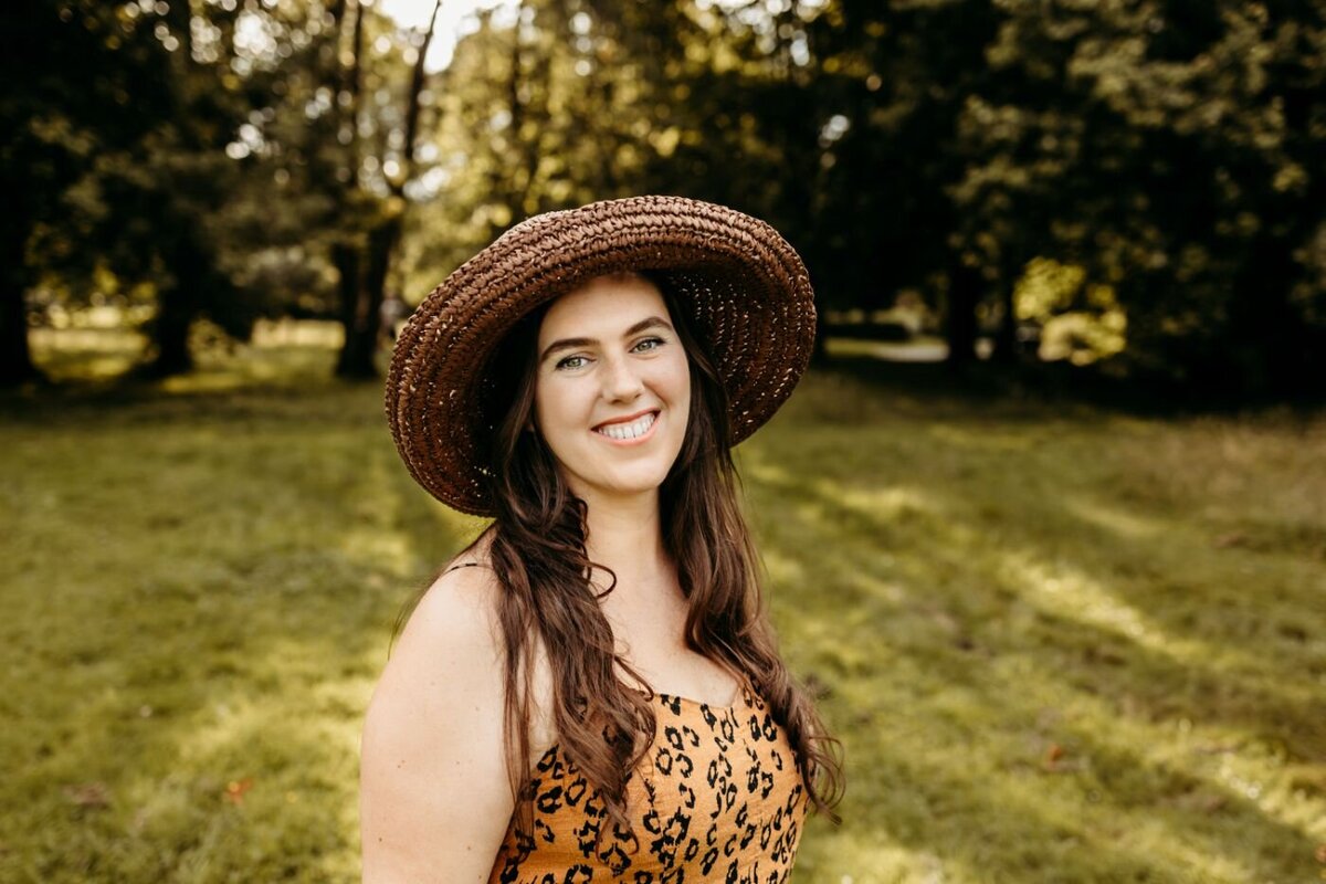 Smiling woman with long brown hair and a wide-brimmed straw hat, wearing a leopard-print top, outdoors on a sunny day.