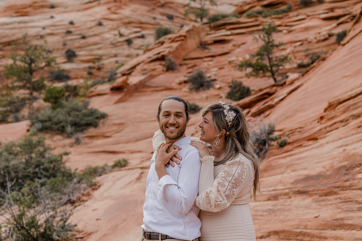 A bride stands behind her groom embracing him on their wedding day. They are surrounded by the red rocks of Utah