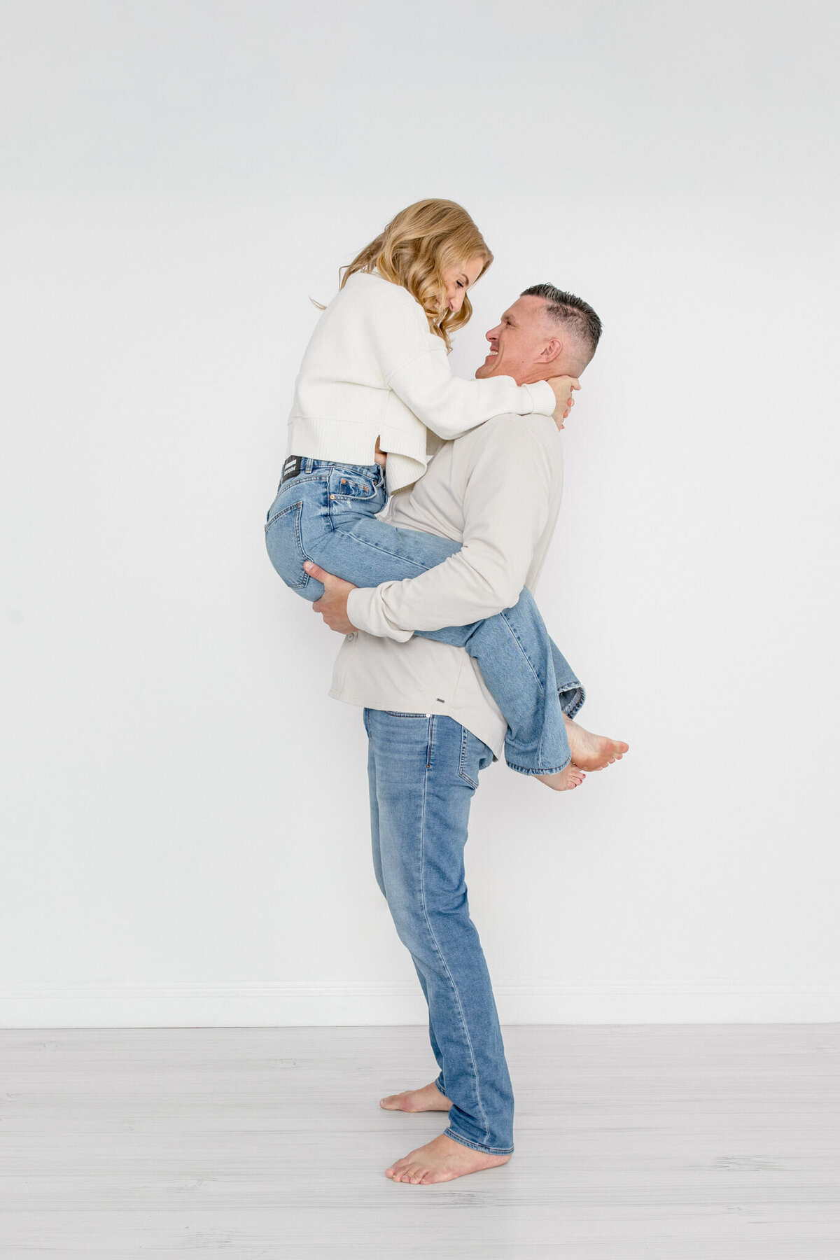 A husband wearing jeans and a white sweater lifting up his wife, whose legs are wrapped around his waist, wearing jeans and a white sweater, hugging his neck as they face each other with their heads together, laughing.