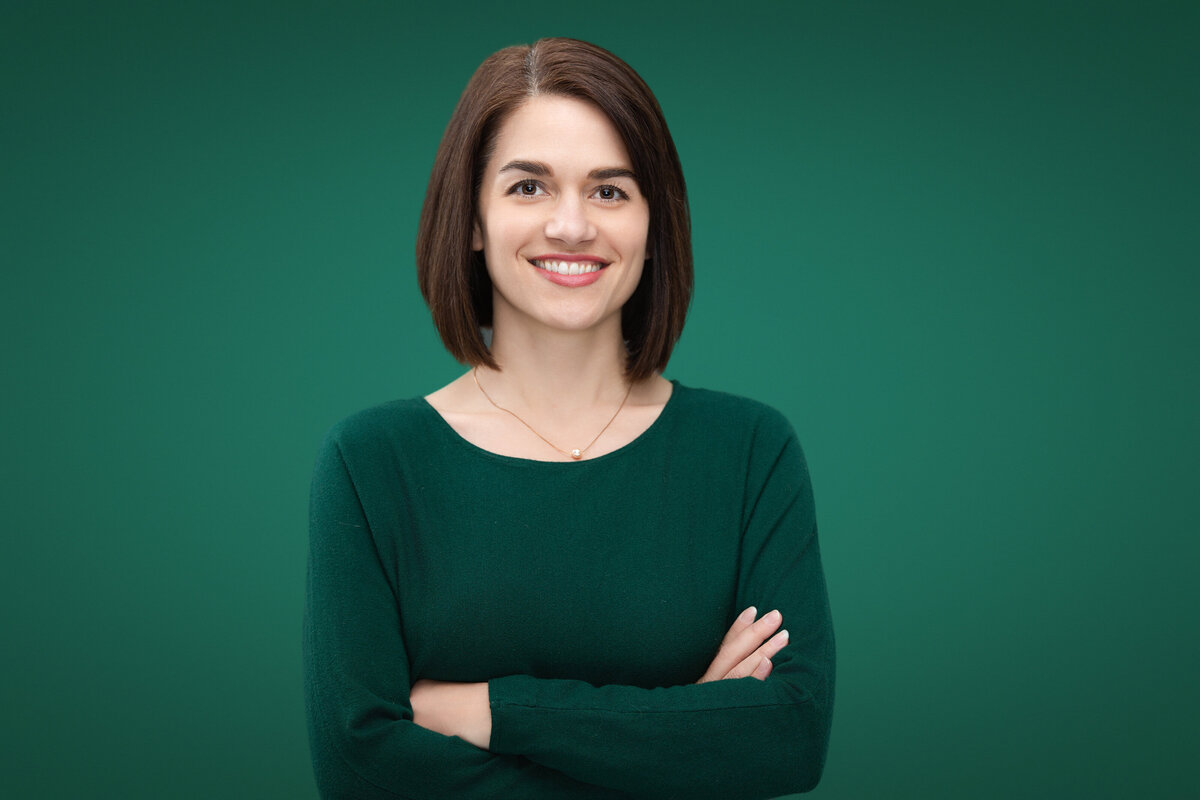 Professional headshot of Krista Brooks, a Caucasian woman, captured against a green backdrop at a Cincinnati studio. Krista is wearing a green sweater, smiling warmly with her arms crossed, ideal for personal branding and corporate profiles. Highlight your professional image with expert headshot photography in Cincinnati.