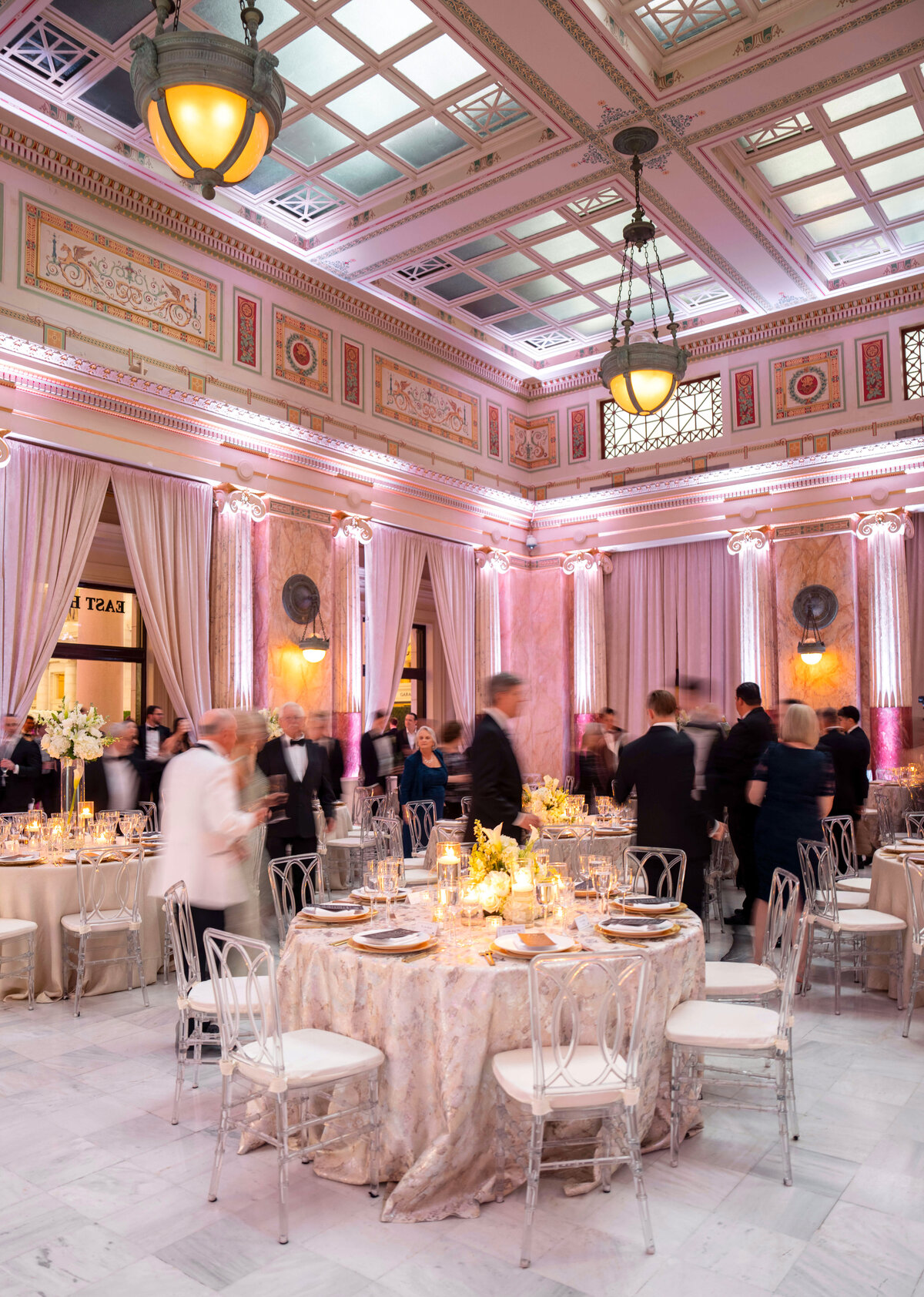 A beautifully decorated ballroom features elegantly set round tables with white floral centerpieces and golden accents. Guests in formal attire mingle under ornate chandeliers and soft ambient lighting, highlighting the rooms classical architecture.