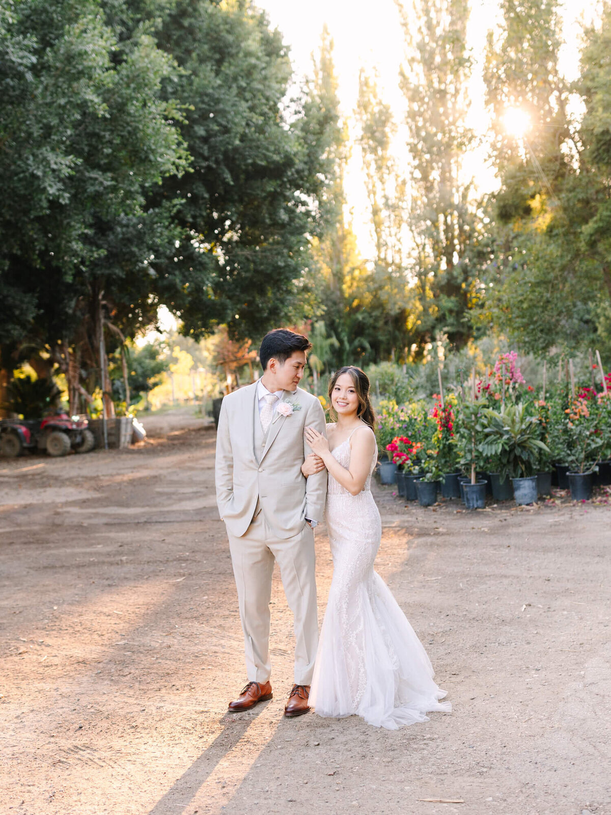 A smiling couple in wedding attire walking hand in hand on a sunlit path with trees in the background.