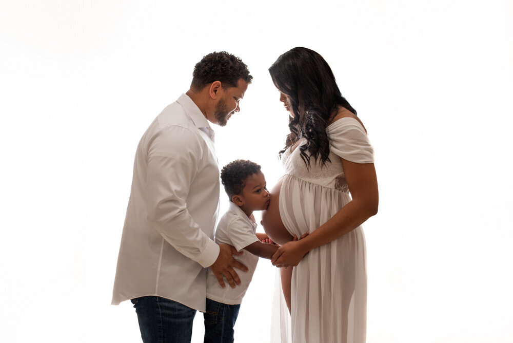 Maternity session with Atlanta, Georgia maternity photographer pregnant woman holding baby bump wearing white sheer maternity  dress on white backdrop posing with husband and 2 year old son