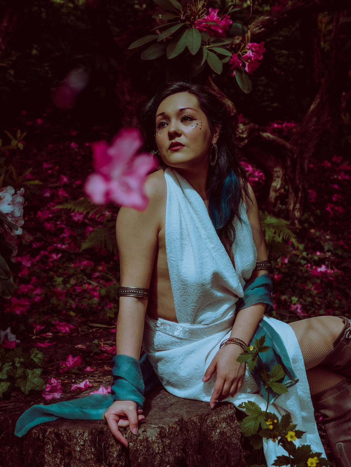 Woman styled like a forest goddess in a white wrap dress surrounded by vibrant pink petals – mystical creative shoot by Mewphotovideo