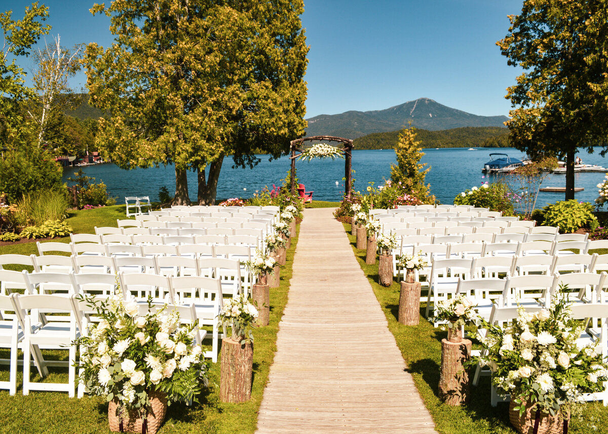 A wedding ceremony is set overlooking a beautiful lake on a green grass garden with white wooden chairs.