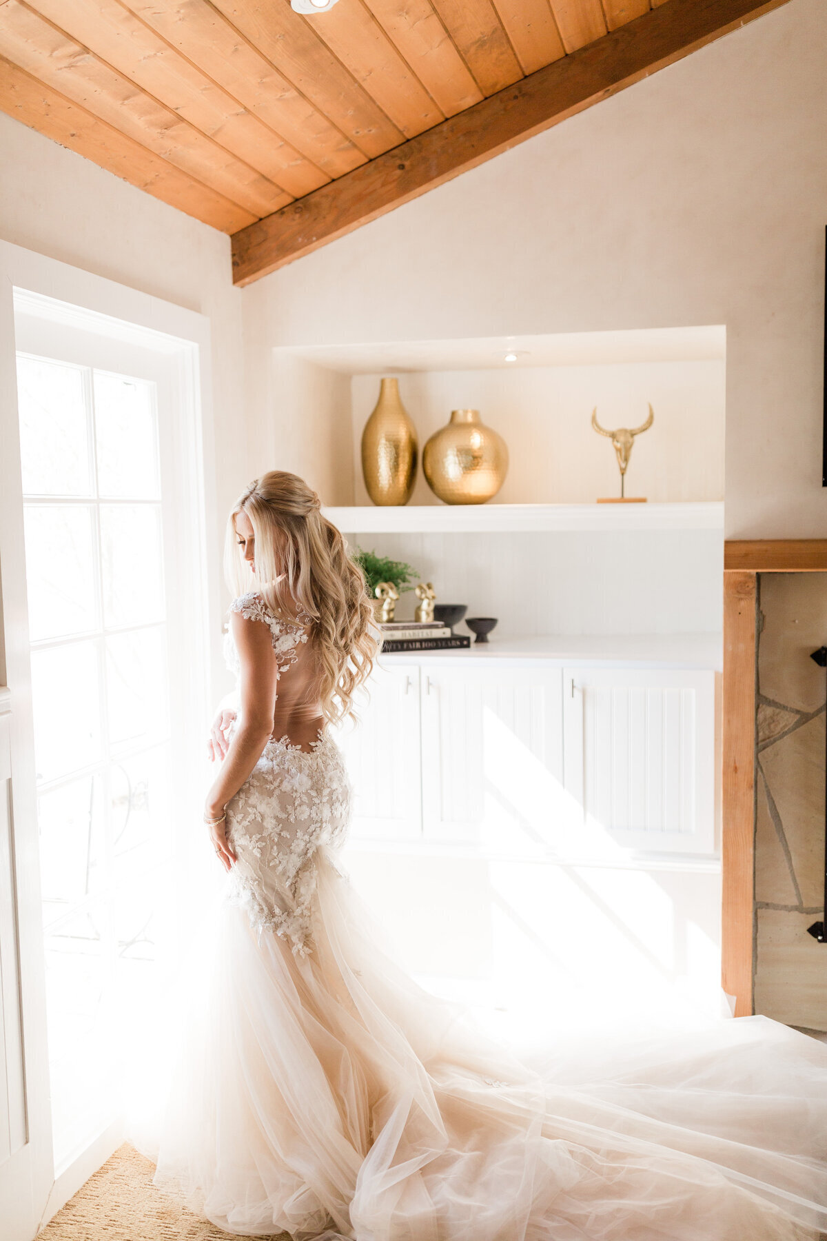 Kohnur Romantic bride in a detailed mermaid gown with natural makeup and long loose waves in a half-updo, posing in a modern farmhouse interior with wood beams.