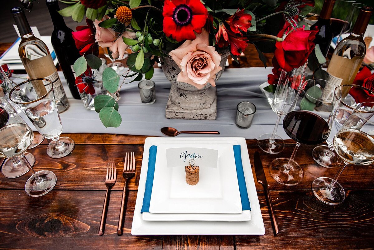 A dark wood farm table set with stacked square white plates wrapped in a teal napkin and topped with a seating card on a champagne cork. On either side of the plate is copper flaatware as well as flat bottomed stemmed wine glasses. Bottles of red and white wine from Arrington Vineyard is set on either side of the large floral arrangement centerpiece. The centerpiece, on a footed marble compote is filled with blush roses, red roses, red poppies, and eucalyptus.