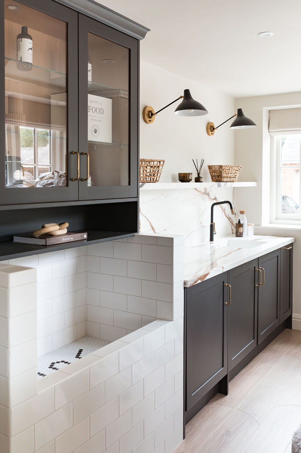 A modern dog wash room with black cabinets, a large white subway tiled washing station and white marble countertops. A black built-in shelf sits above the washing station. A shelf with wicker baskets sits above the counter, along with two black lights.