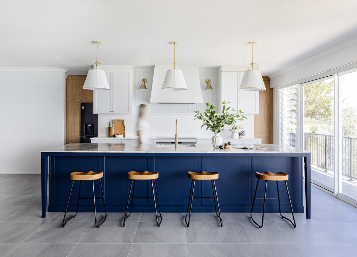Modern kitchen designed by a Newcastle Interior Designer, featuring a large blue island, wooden stools, white cabinets, pendant lights, and a person walking by.