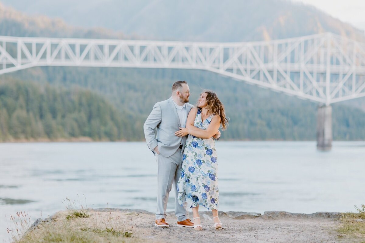 A happy couple in blue stand on the edge of a river with a white bridge backdrop