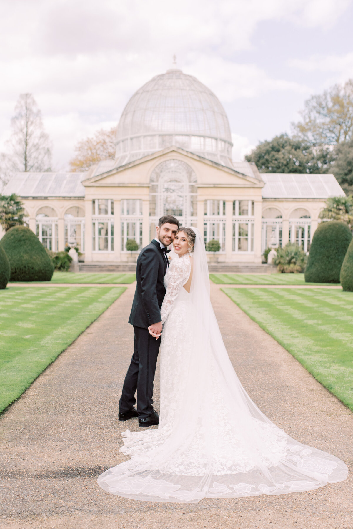 Bride and groom are standing side by side looking back at the camera, while holding hands, Syon Park is in the back ground of the image. The bride is wearing a white lace dress and veil and the groom is in a black tux