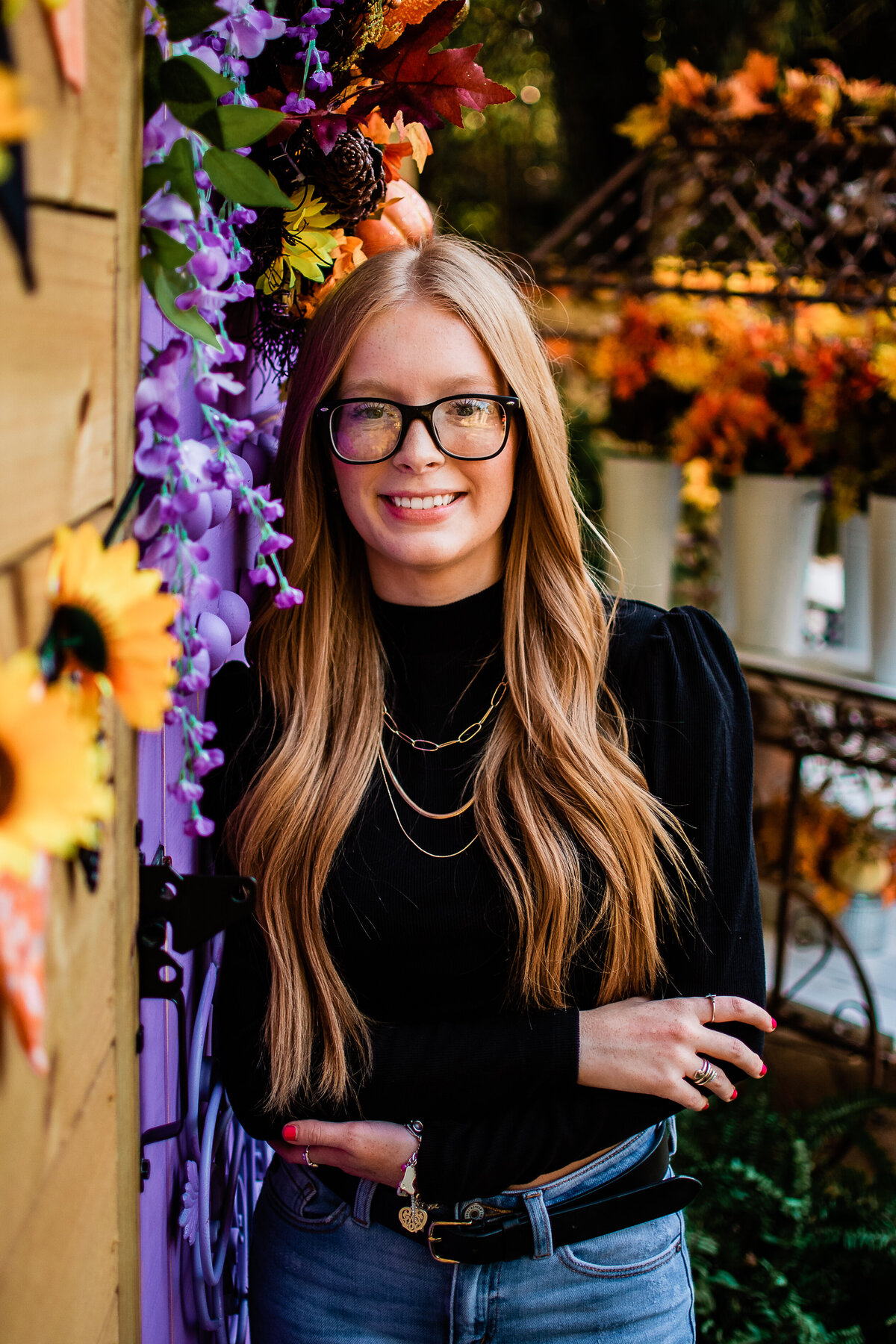 A senior wearing a black shirt and gold necklace leans against a door decorated with fall themed flowers.