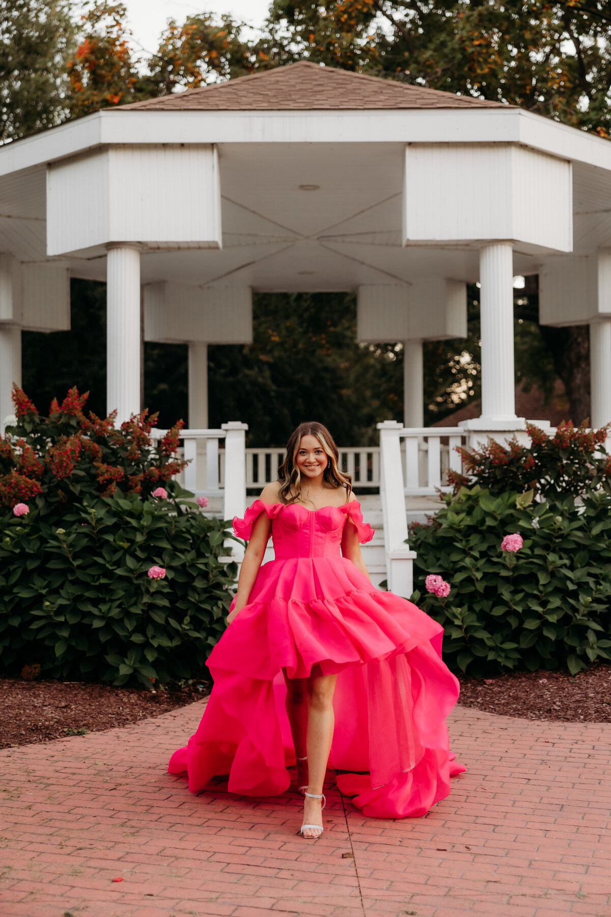 A girl in a hot pink dress does a runway walk in front of a gazebo.