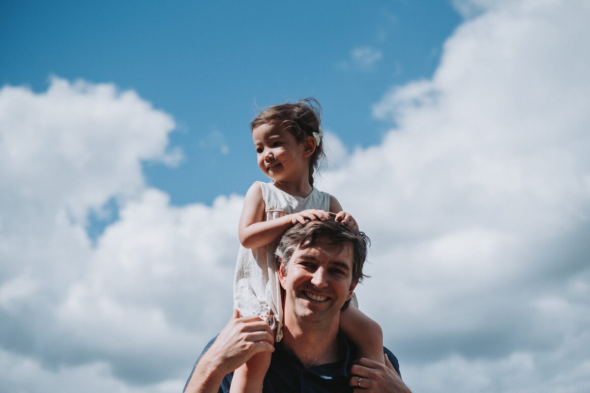 Father carries toddler daughter on his shoulders with sunny blue sky in the background