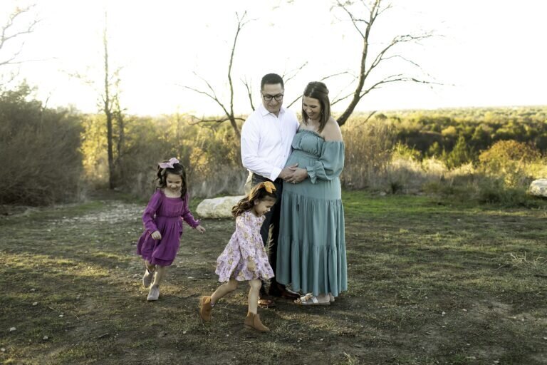 A joyful maternity photo of the expectant mom and dad standing together in the park, with their two young girls playfully running around them, creating a lively and loving family moment, captured by Chunky Monkey Photography.