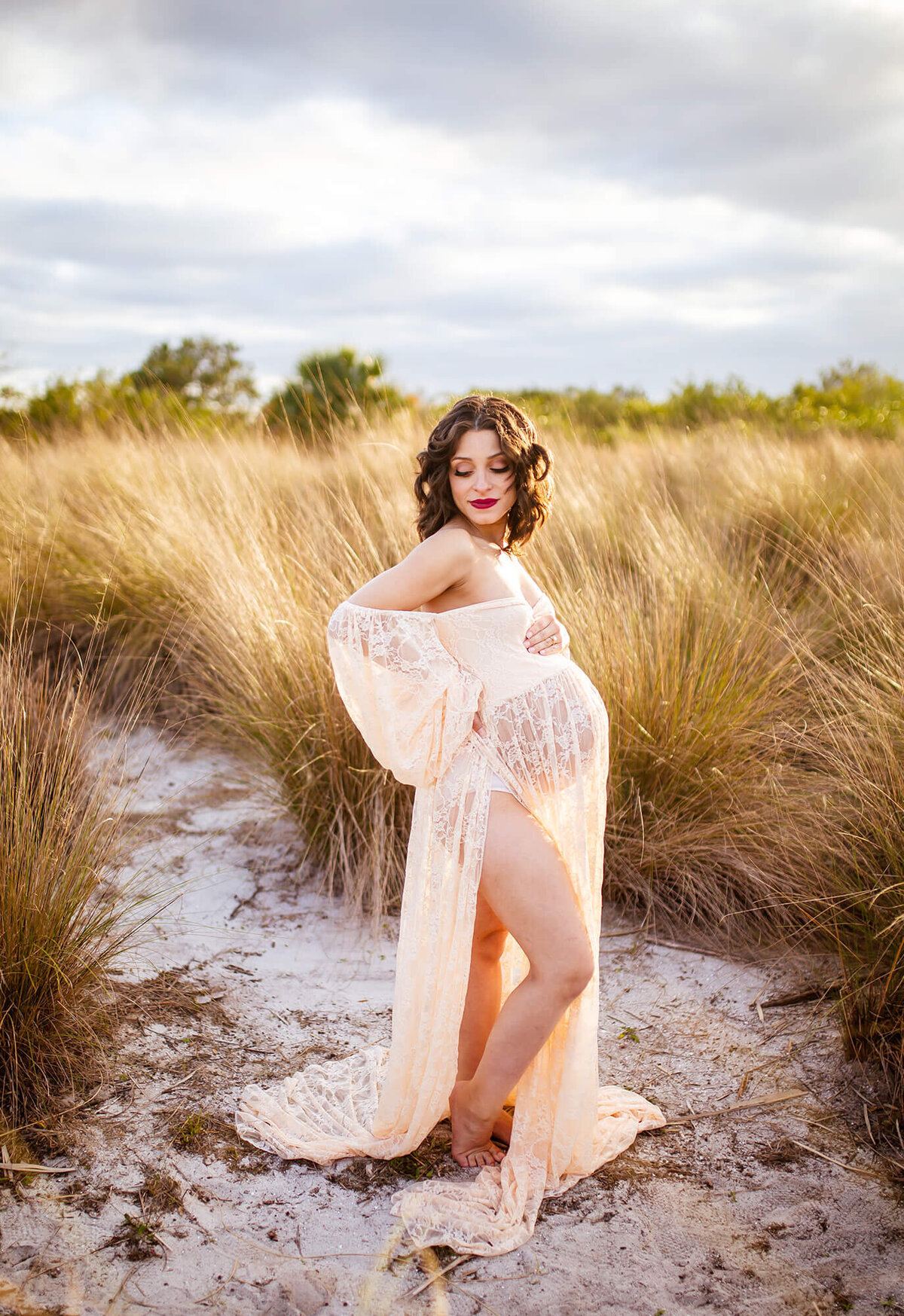 Golden light sunset maternity session. Woman is holding belly and looking down in lace dress. Captured by Sarah Wyndham Photography at Cypress Point Park