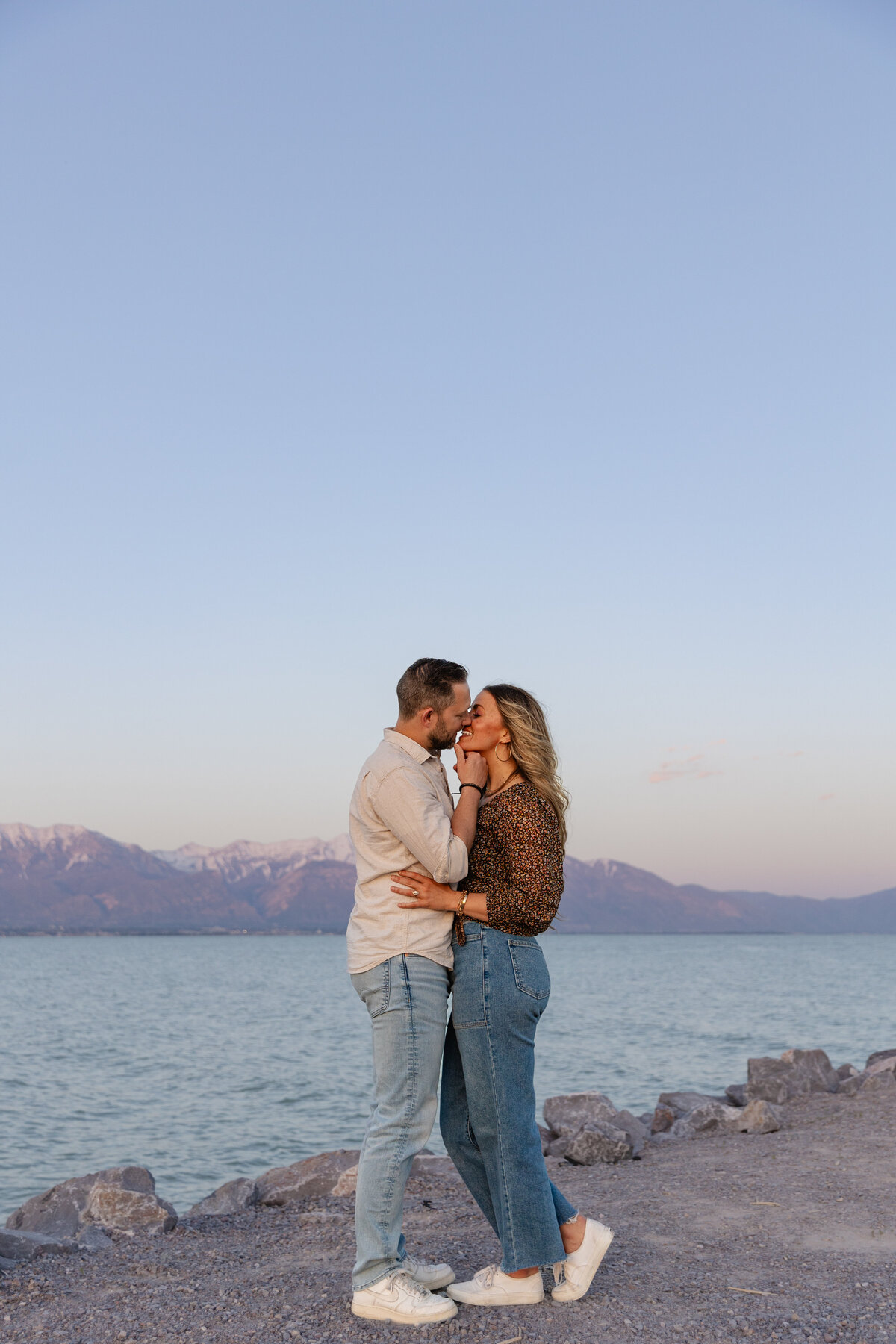 A couple shares a gentle kiss on the rocky shore of Utah Lake as the sun sets behind the mountains. The peaceful blue water reflects the fading daylight.