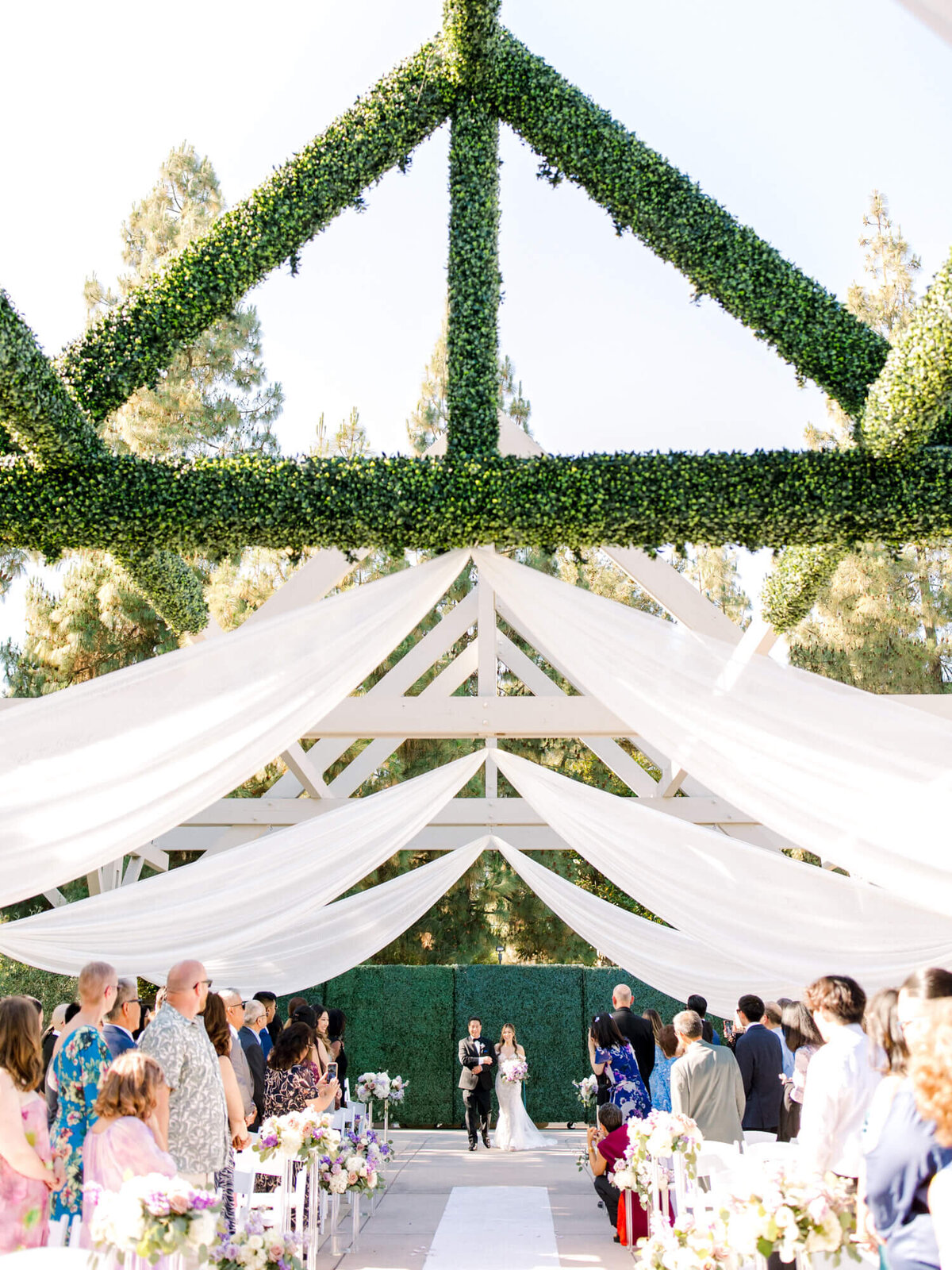 A bride and her father walk down an aisle adorned with flowers under a trellis draped with white fabric and greenery, surrounded by guests and trees, creating a serene, joyful atmosphere.