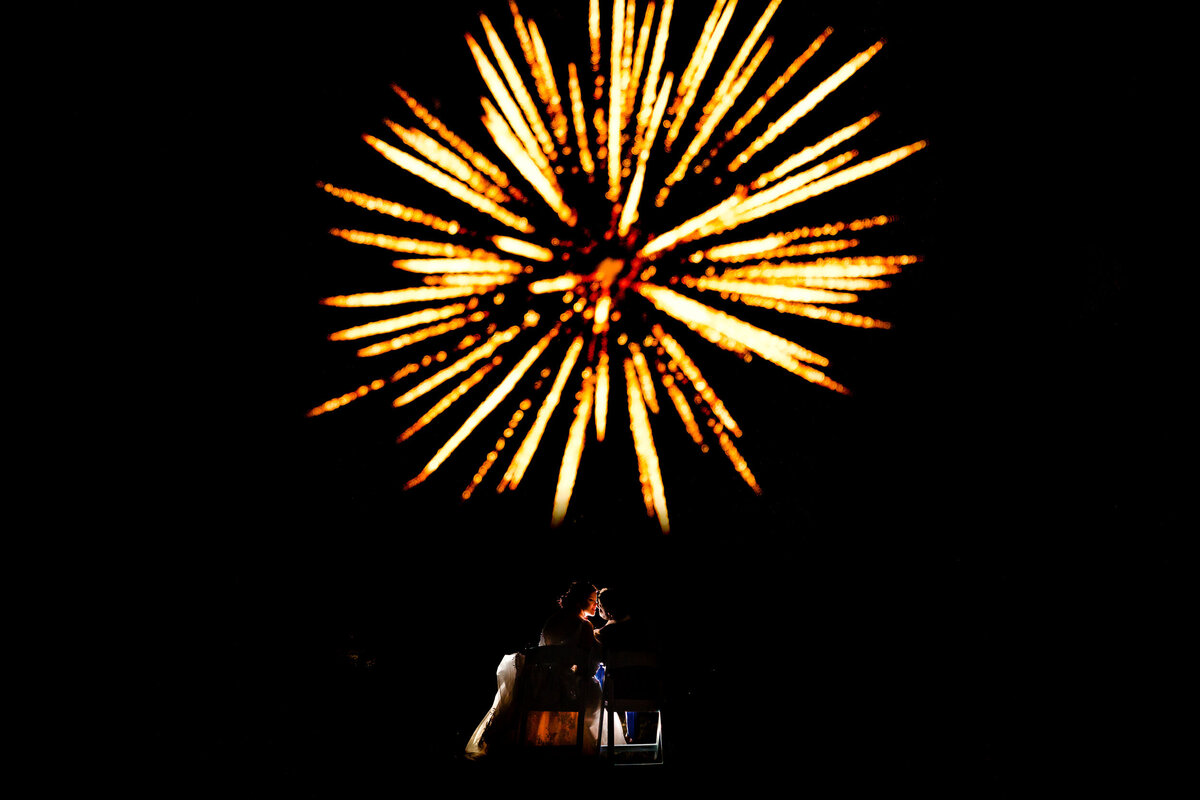 One of the top wedding photos of 2020. Taken by Adore Wedding Photography- Toledo, Ohio Wedding Photographers. This photo is of a bride and groom kissing in the dark as a massive orange and yellow  firework explodes over head