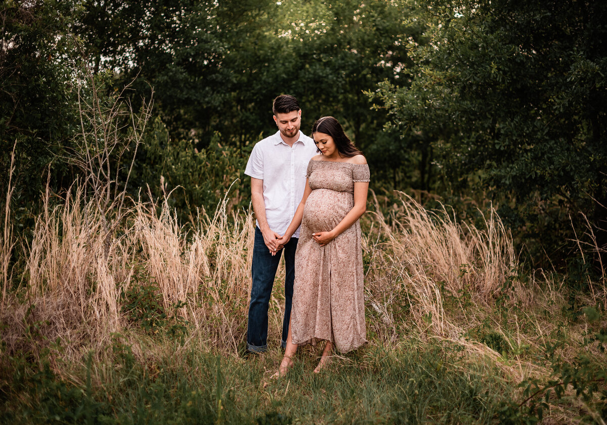A mom to be holds her boyfriend's hand while they both look at her belly in a field of long grass.