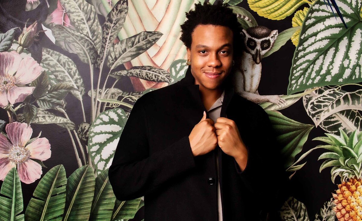 Male musician portrait Justin Lewis holding black overcoat lapels with tropical wallpaper background