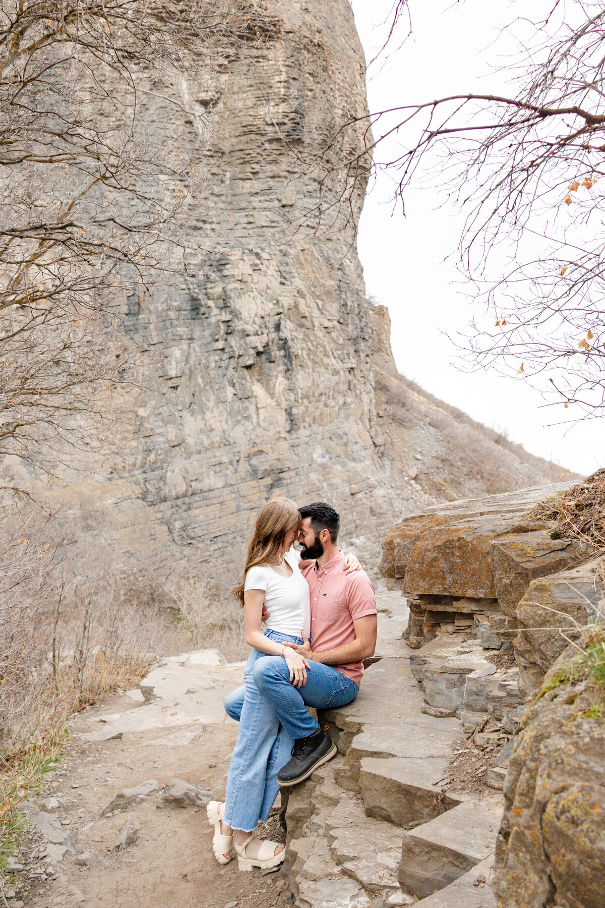 Engaged couple sitting close on a rocky trail in Orem, Utah, sharing an intimate moment with a dramatic cliffside in the background.