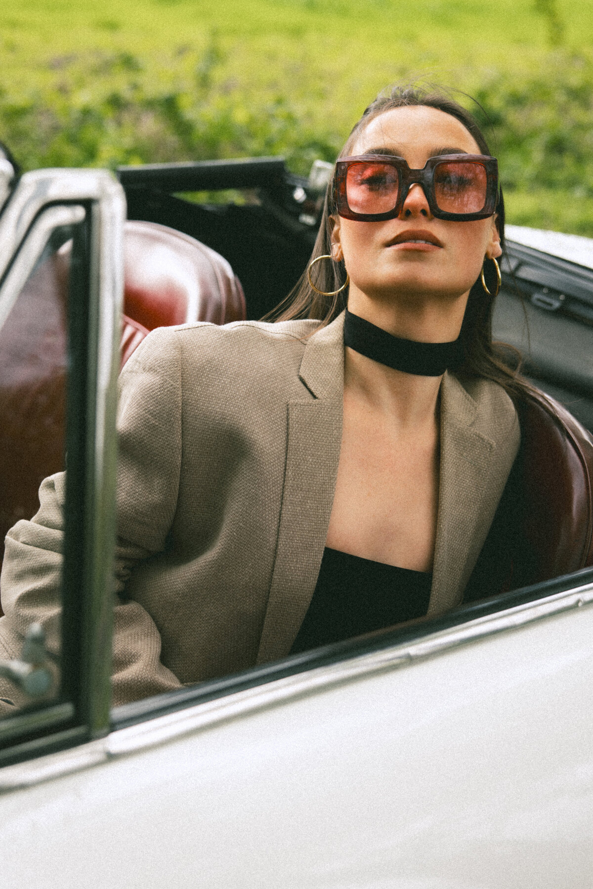 The model poses with oversized sunglasses and a tailored blazer, embracing a chic and timeless look. Her relaxed posture contrasts with the structured lines of the car, creating a harmonious blend of elegance and attitude. Photographed by Bo Eppink.