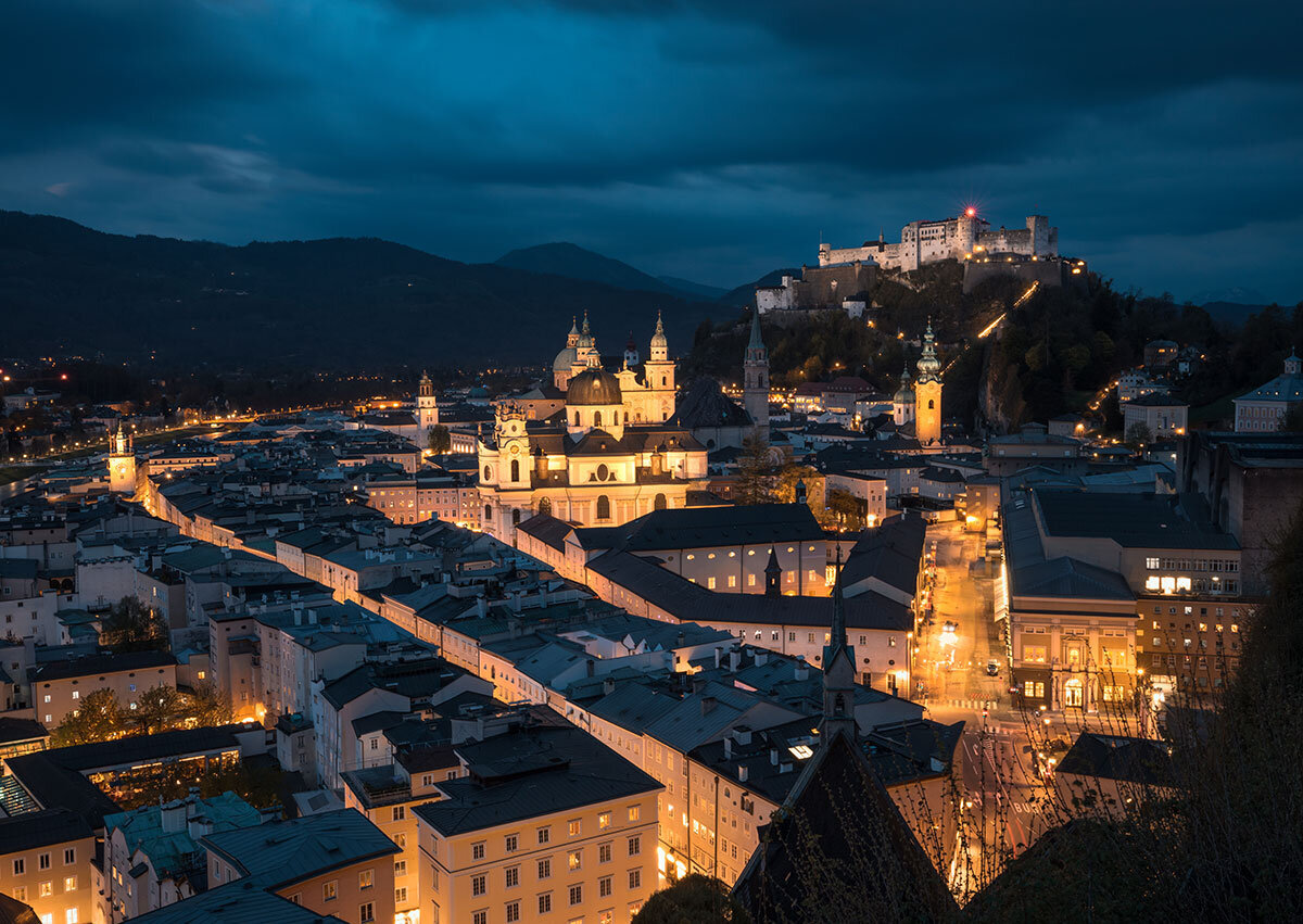 Ein Nachtwächter in traditioneller Kleidung mit Hut und Laterne führt eine Gruppe von Teilnehmern durch die beleuchteten, historischen Gassen der Salzburger Altstadt. Die nächtliche Atmosphäre sorgt für eine mystische Stimmung.