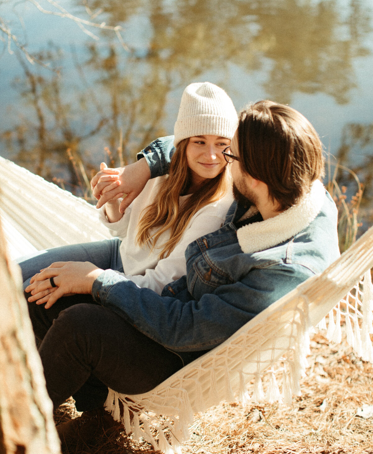 A couple is wearing a winter outfit and sitting in a hammock together with a lake behind them.
