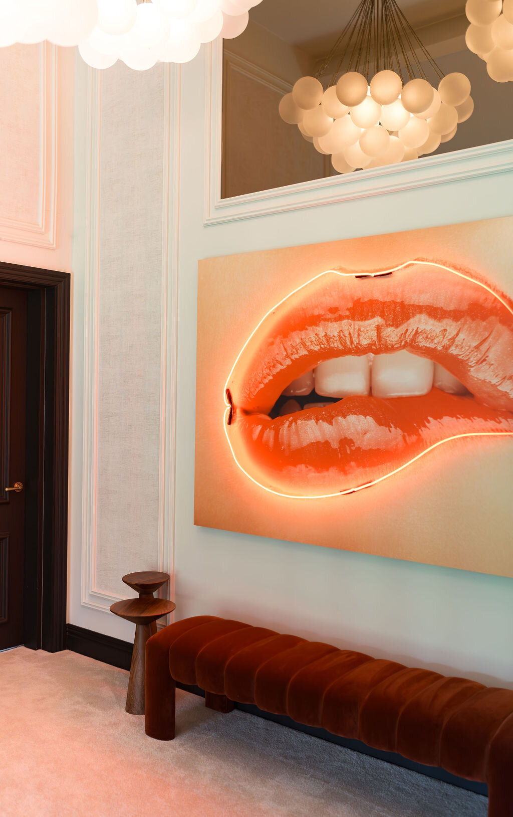 A large, vibrant painting of red lips biting the lower lip is displayed on a white wall. The painting is illuminated with LEDs, casting a soft pink light on the room. Below the painting is a cushioned, burnt orange velvet bench flanked by two wooden side tables. The room features a modern and minimalistic design with neutral tones.