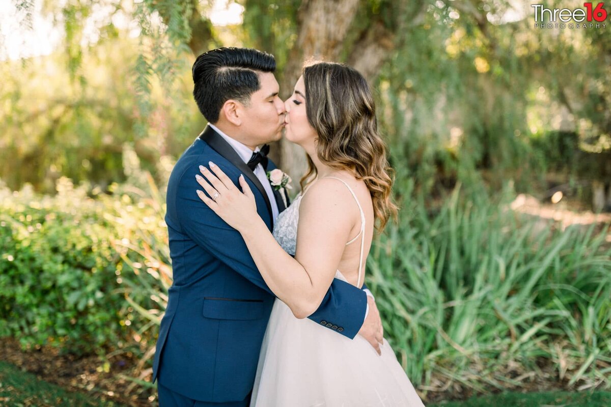 Tender kiss for newly married couple in front of green bushes