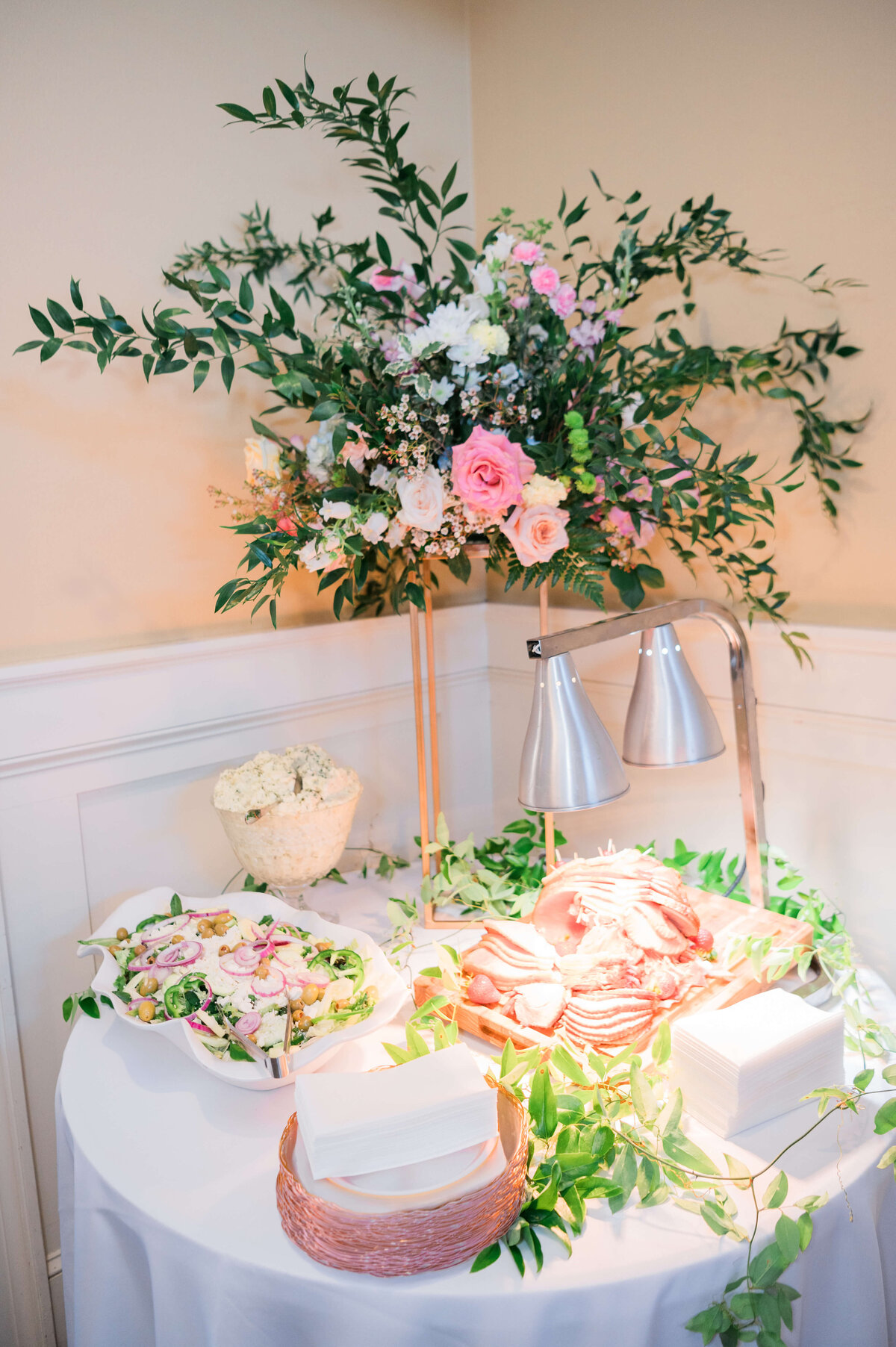 A round table with a white tablecloth holds a floral arrangement in pink and white, a salad bowl, a dish of sliced meats under heat lamps, stacked napkins, and greenery decor.