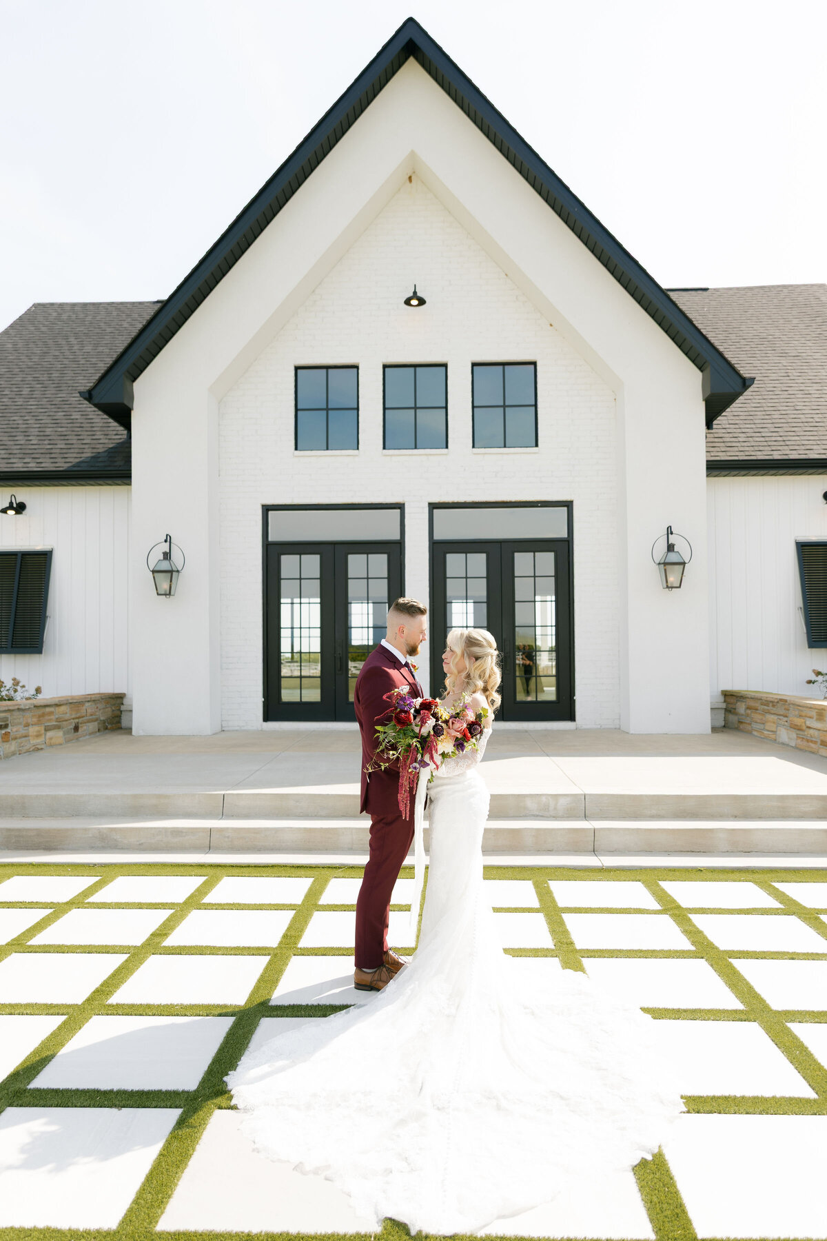 The bride and groom share a quiet moment standing in front of a modern white wedding chapel with black-trim windows. The bride wears an off-the-shoulder lace gown with a long train while holding a lush fall-colored bouquet, and the groom wears a burgundy suit.