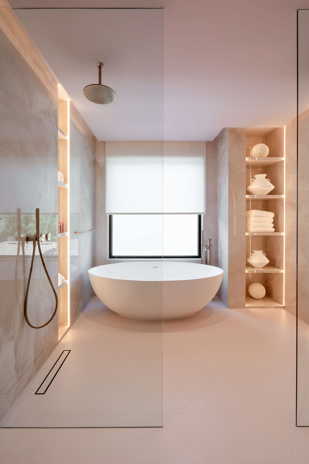 Spacious en suite bathroom with a  chic light pink aesthetic. The walls are a light pink marble. There is a large soaking tub against the far wall, and open showers with rose gold hardware on the left.