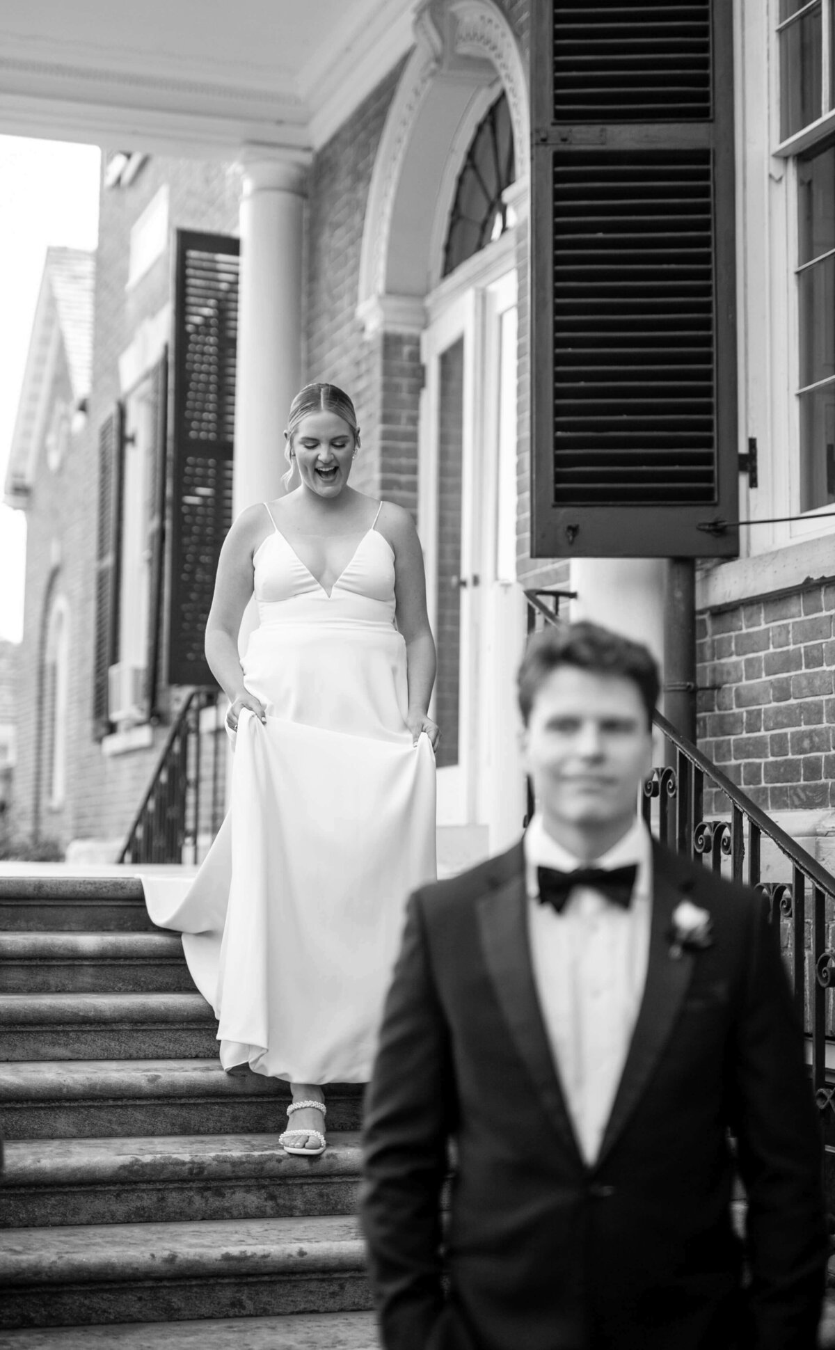 A bride in a white gown excitedly descends steps towards a groom in a suit, who stands in the foreground. They are on the porch of a brick building with large windows and shutters. The scene conveys anticipation and joy. Black and white photo.