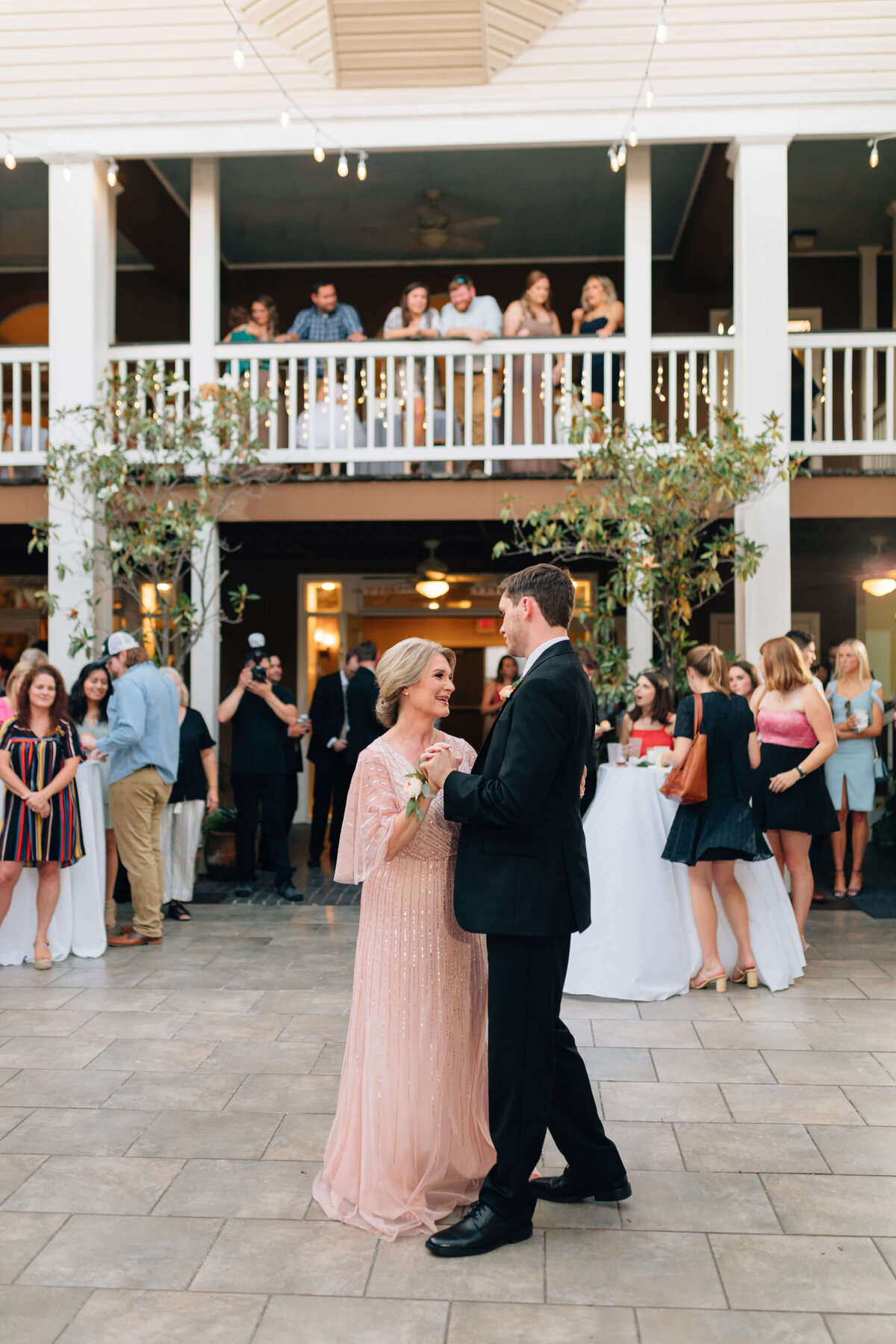 A woman in a blush pink dress and a man in a black suit dance together outdoors while guests watch and mingle. Onlookers stand on a balcony above, under string lights.