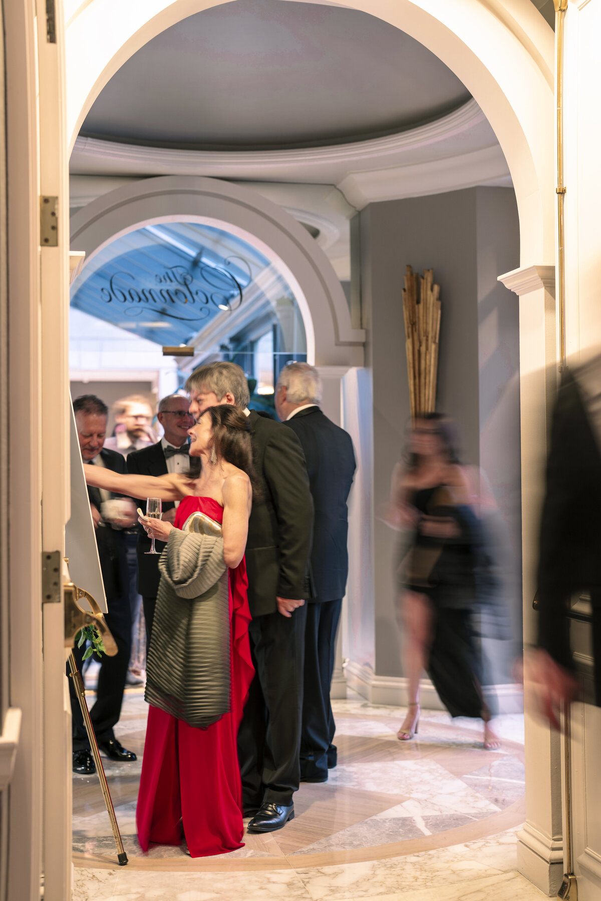 A group of elegantly dressed people gather in a luxurious hallway. A woman in a red gown, holding a drink, stands out in the foreground. The scene includes an arched entrance and blurred figures suggesting movement, creating a lively atmosphere.