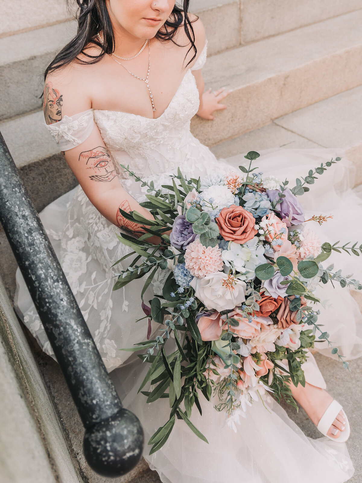candid photo of a bride wearing an off-the-shoulder white and champagne floral lace wedding gown sitting on marble stairs holding a cascading red, pink, peach, white, blue and purple wedding bouquet taken by Massachusetts Wedding Photographer Avid Aperture Photography located at Customs House Square in New Bedford Massachusetts
