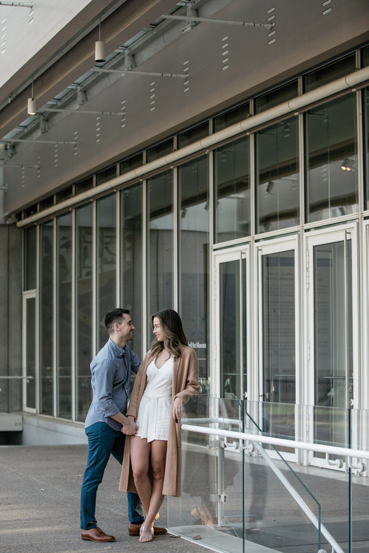 Engaged couple at the Kimbell Art Museum in Fort Worth, she leaning on a railing while he stands in front, sharing a playful, flirty moment