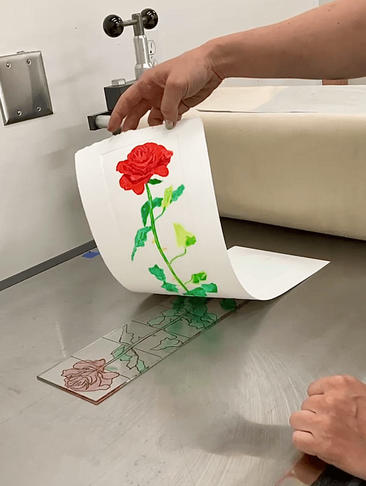 A hand pulls a bold red rose print from etched plexiglass on the press, highlighting the crisp linework and soft watercolor effect.