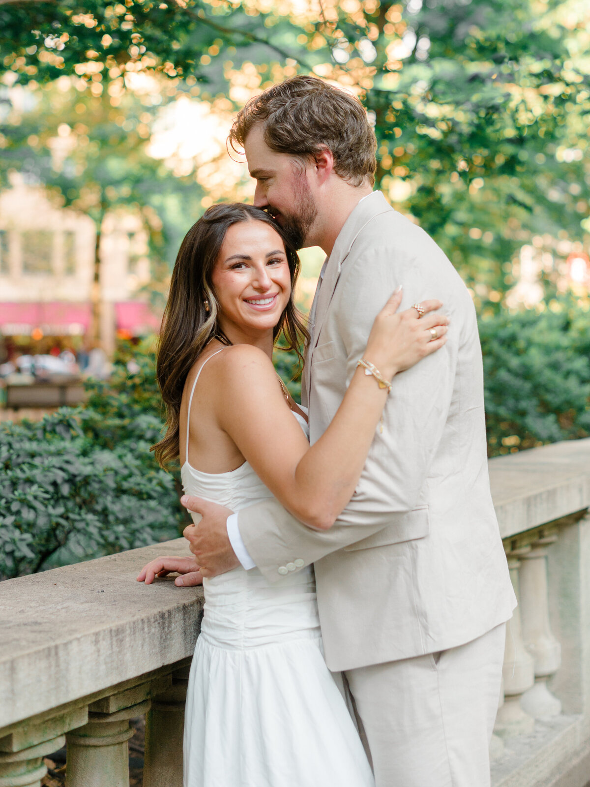 A joyful bride-to-be smiles at the camera as her fiancé gently kisses her forehead during a quiet moment in Rittenhouse Square. Surrounded by soft greenery and warm golden light, the couple embraces against a historic stone railing. Captured by Elizabeth Kane Photography, a Luxury Philadelphia Wedding Photographer.