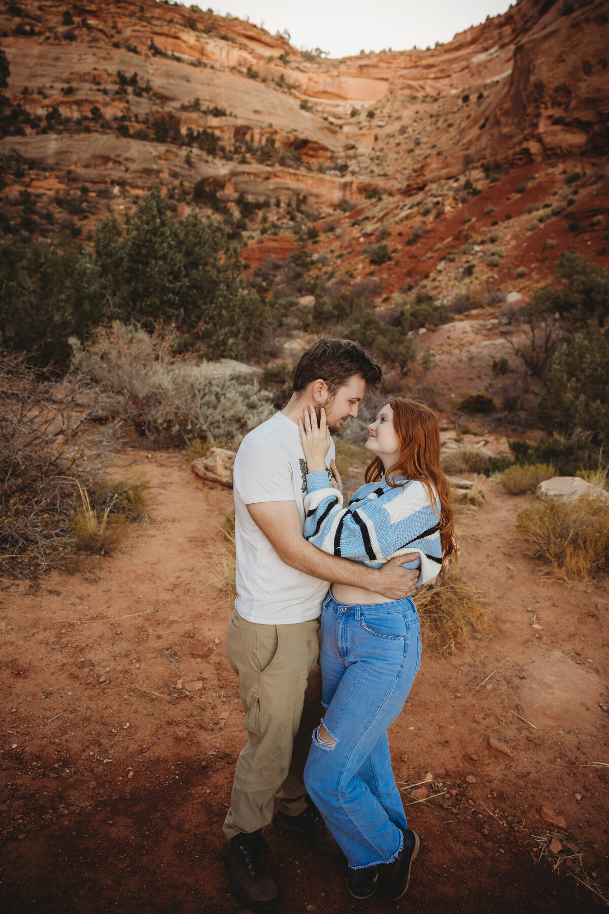 A couple wraps their arms around each other and stare at each other lovingly amidst the red rocks of the landscape.