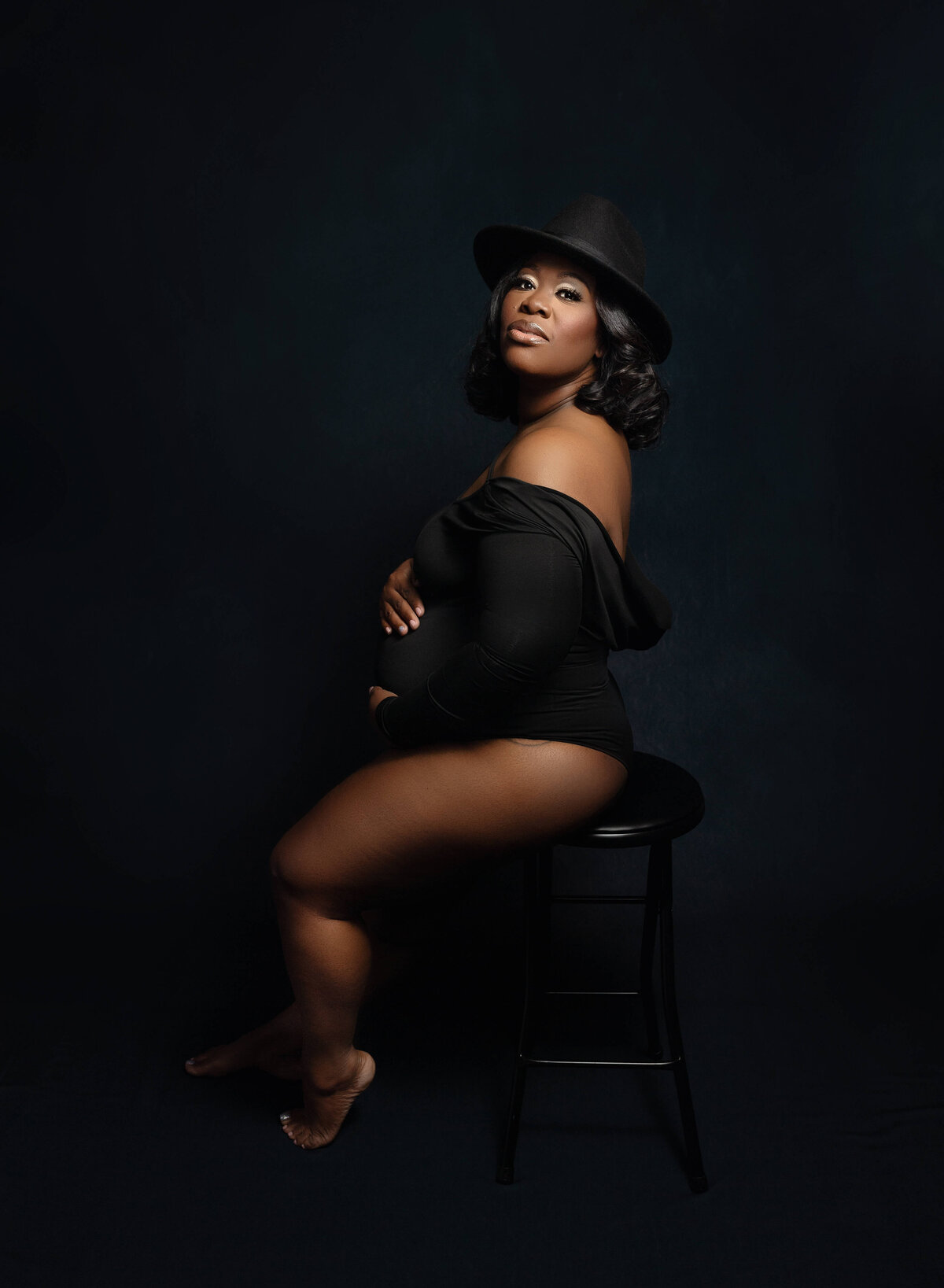A black mother to be sits on a stool in a black one-piece maternity suit and a black hat in a New Orleans Maternity Photographer studio