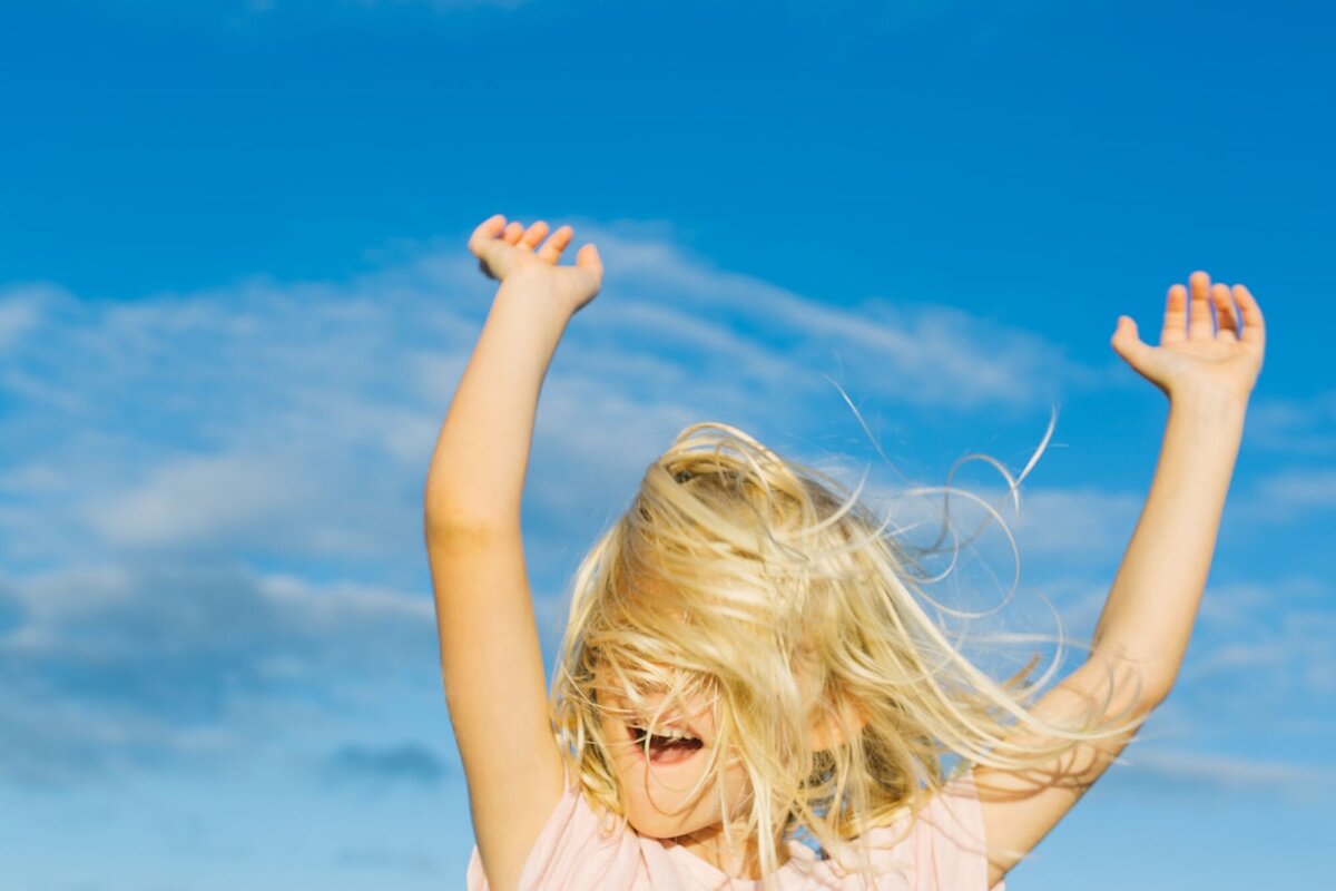 A little girl throws her arms up in the air as the wind flushes the hair in front of her face.