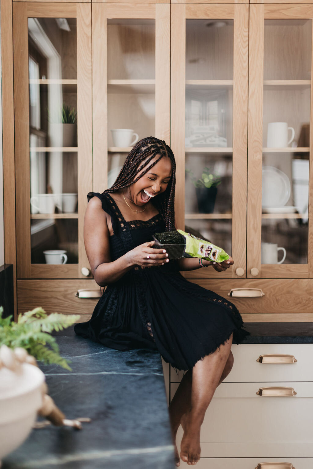 Juma Waganda in a black dress sitting on the kitchen counter holding a snack