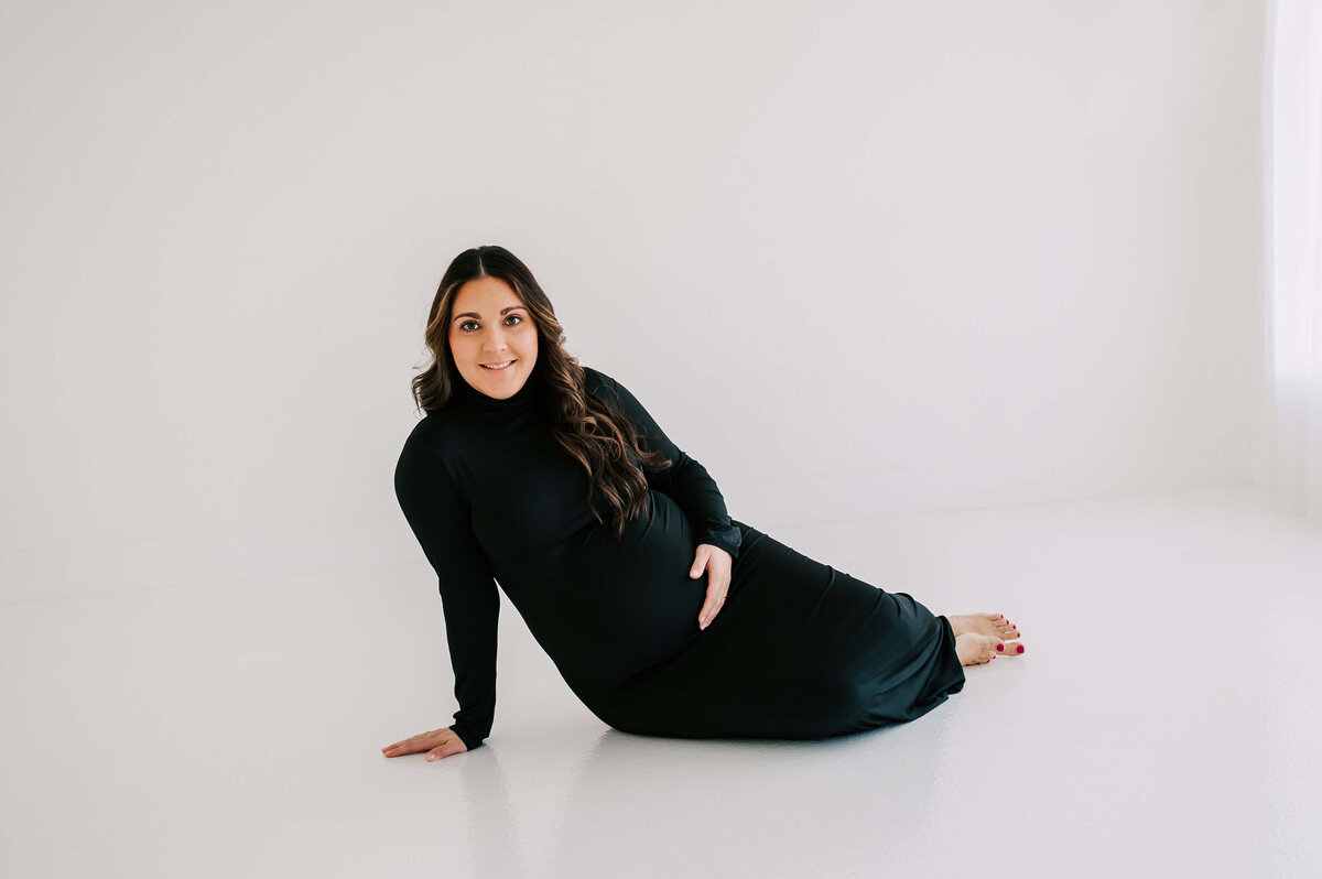 pregnant mom in black dress smiling while sitting on the floor of maternity photography studio in Joplin