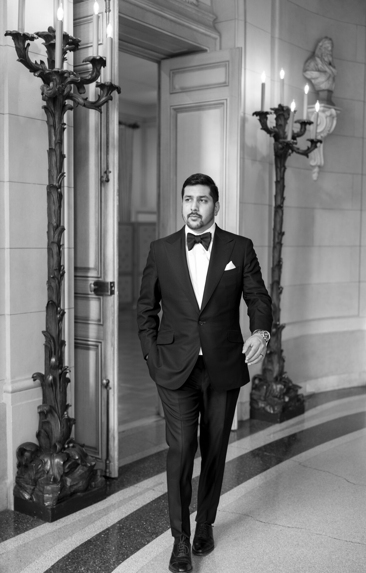 A man in a tuxedo walks confidently through an elegant hallway. The scene is black and white, featuring ornate woodwork, tall candlesticks, and a statue on the wall, evoking a classic, sophisticated atmosphere.