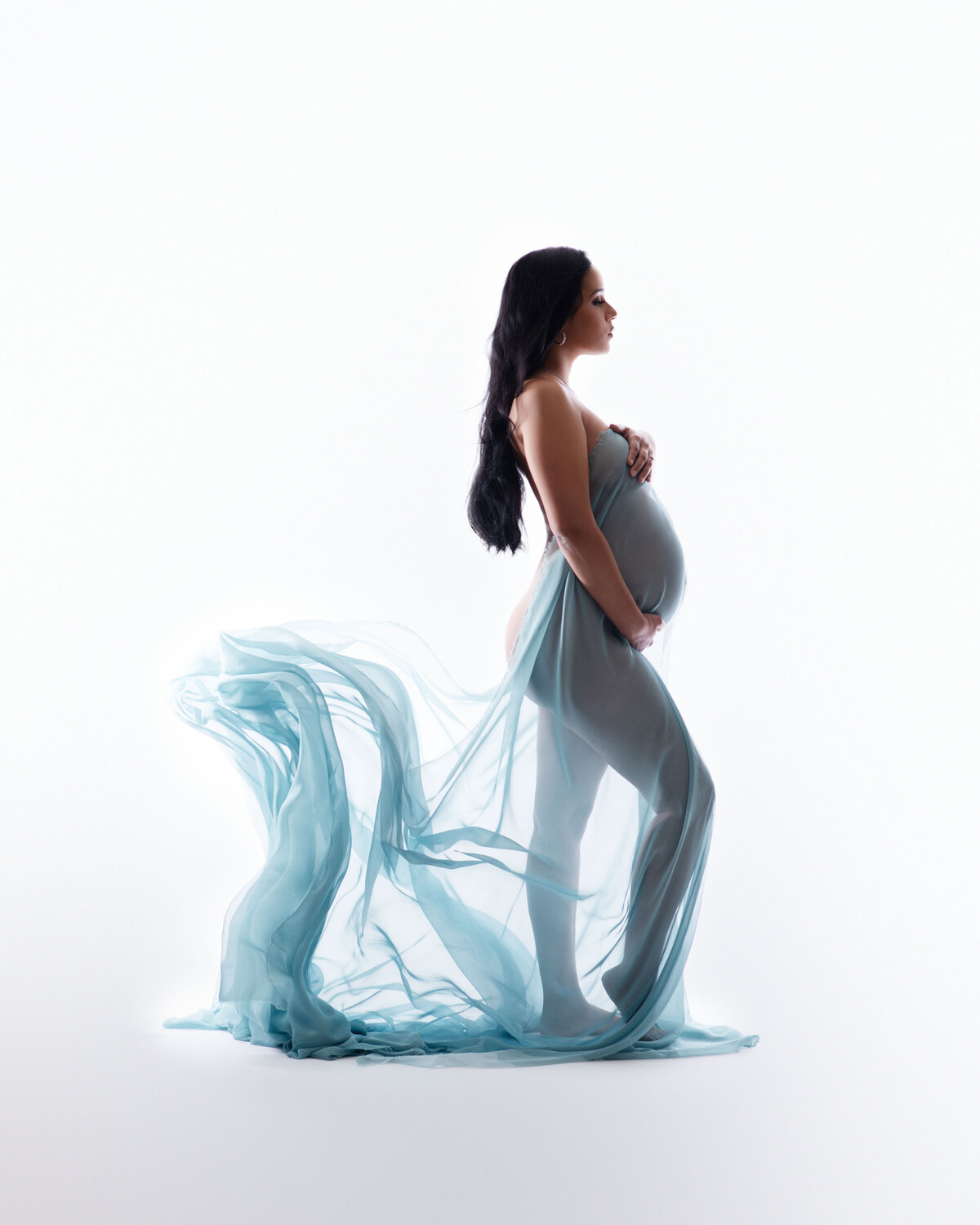 Pregnant long brown haired woman standing to side posing with one leg up holding baby bump and other hand over breast wearing a soft blue flowy fabric