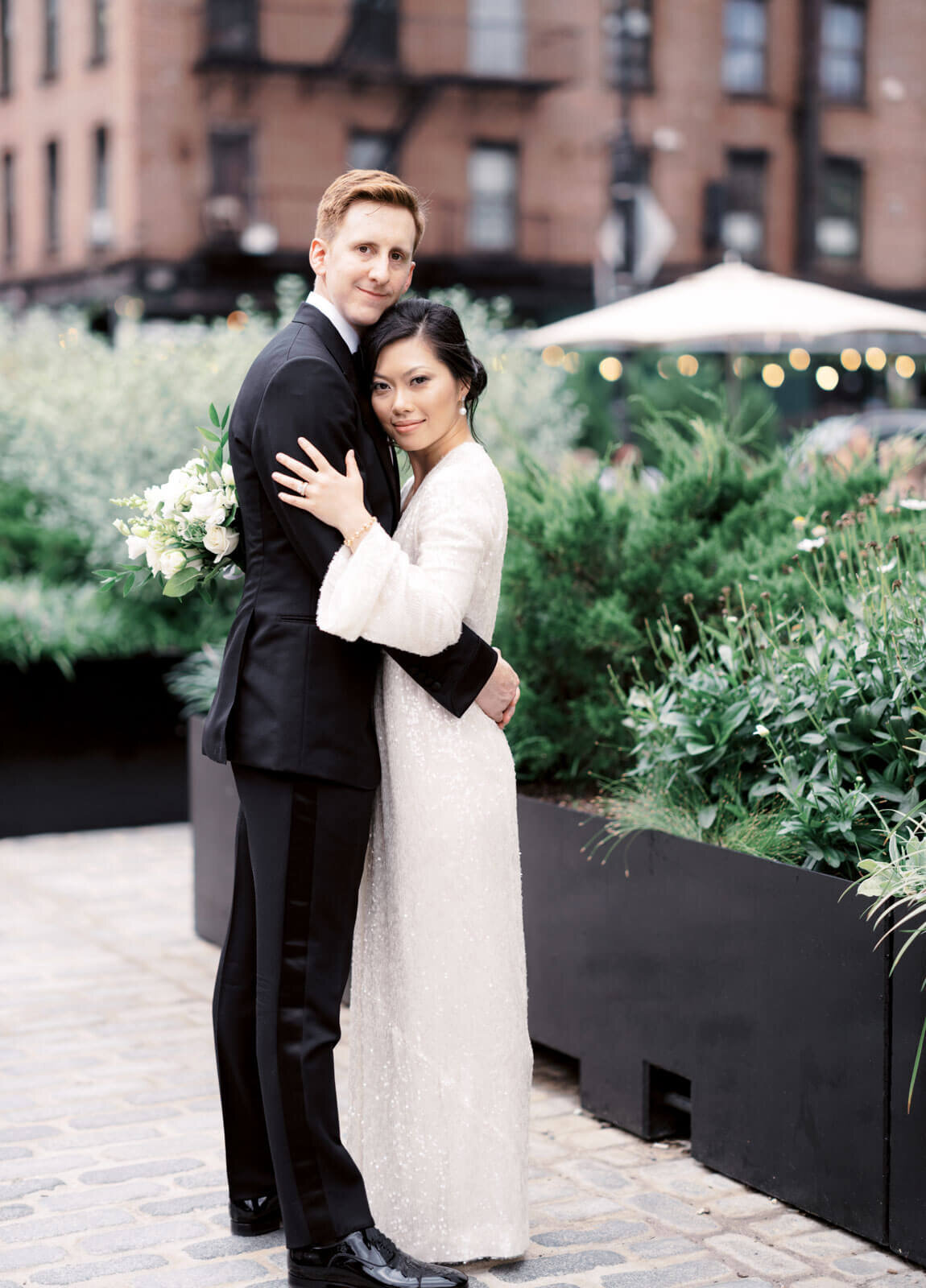 The bride and groom are lightly hugging each other, with some plants and a red brick building in the background in West Village, NYC.
